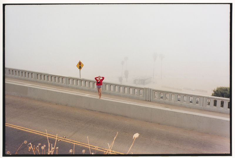 Model in a red top and shorts standing on a concrete bridge overpass in dense coastal fog.