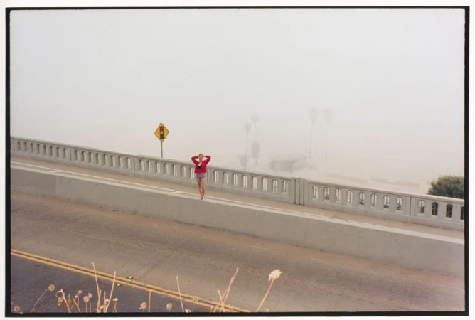 Model in a red top and shorts standing on a concrete bridge overpass in dense coastal fog.