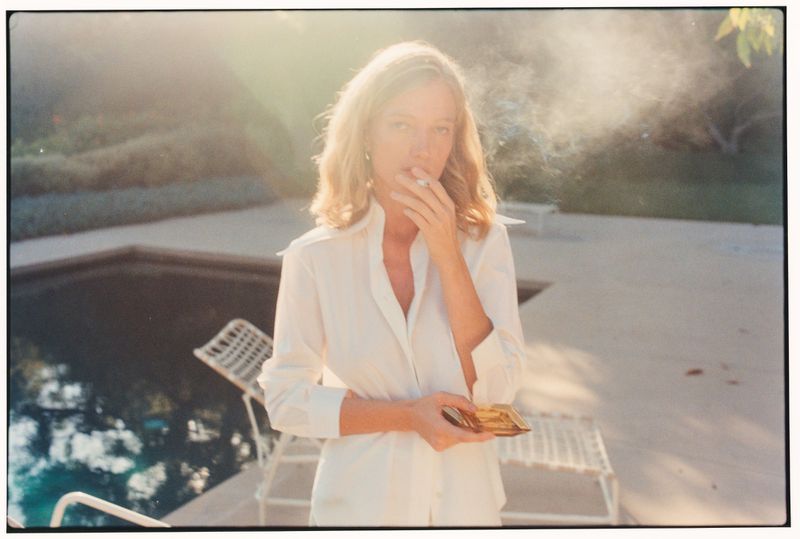Woman in an unbuttoned white shirt standing poolside in sunlight holding a gold compact while smoking with visible smoke drifting.