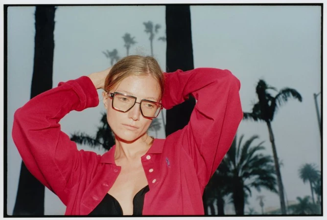 Model in oversized red long-sleeve polo and large square eyeglasses posing outdoors against silhouetted palm trees.