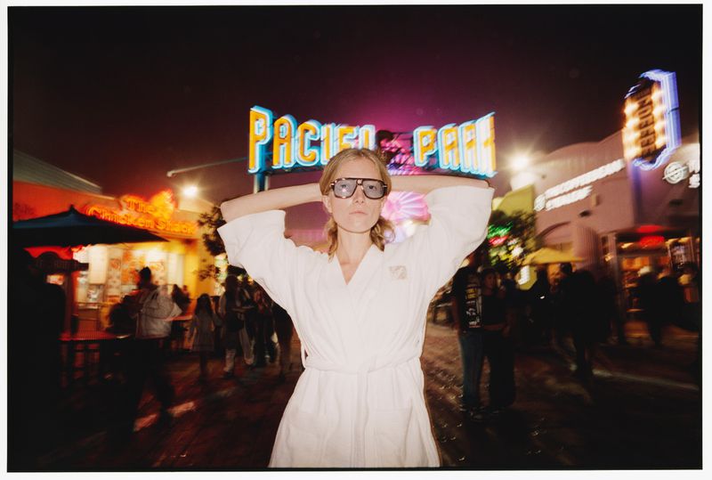 Model in a white bathrobe and oversized sunglasses posing at night in front of the illuminated Pacific Park sign.