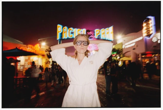 Model in a white bathrobe and oversized sunglasses posing at night in front of the illuminated Pacific Park sign.
