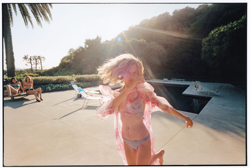 A blonde woman in a bikini and sheer pink coverup runs poolside under bright sunlight while two women sit on deck chairs.