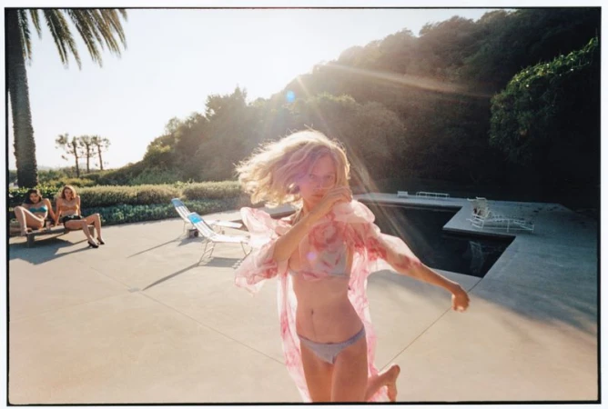 A blonde woman in a bikini and sheer pink coverup runs poolside under bright sunlight while two women sit on deck chairs.