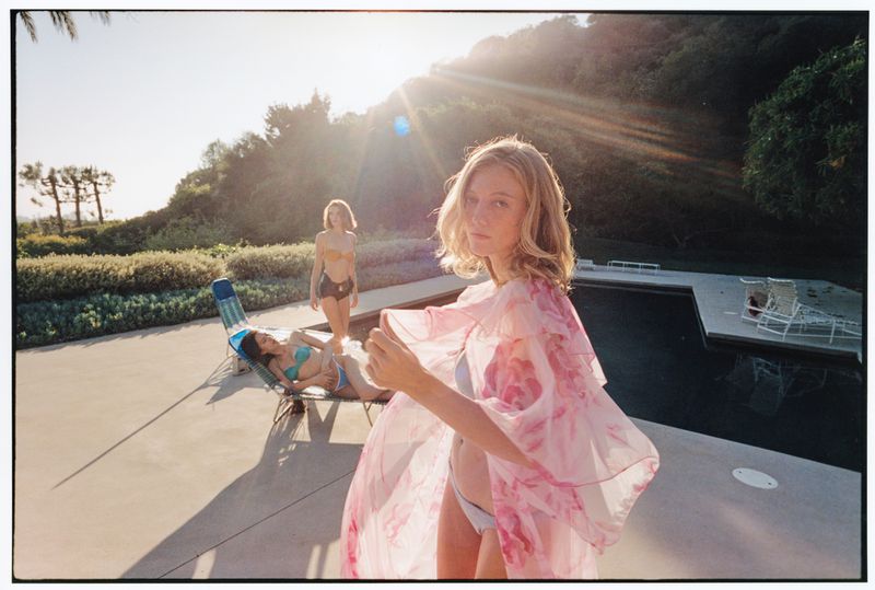 Blonde model in a sheer pink floral robe standing poolside with two models in swimwear under bright sun flare.