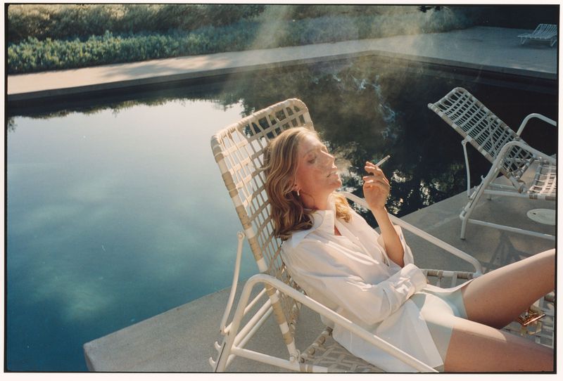 Woman wearing an oversized white shirt smoking a cigarette while relaxing on a white lounge chair beside a swimming pool.