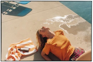 A woman in an orange t-shirt sunbathes beside a swimming pool next to a striped towel and lotion bottle on the concrete deck.