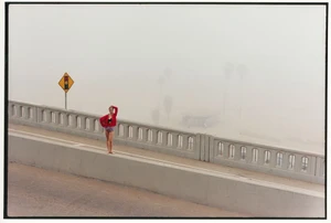 A woman in a red cardigan and patterned shorts stands on a concrete bridge ledge beside a yellow traffic sign in heavy fog.