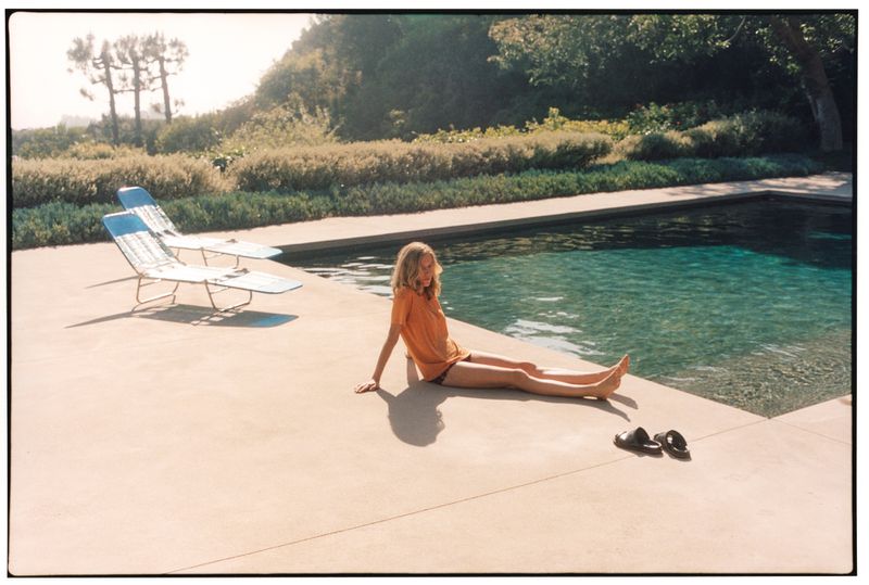 A woman in an orange t-shirt sits poolside on concrete paving next to an outdoor swimming pool and sun loungers.