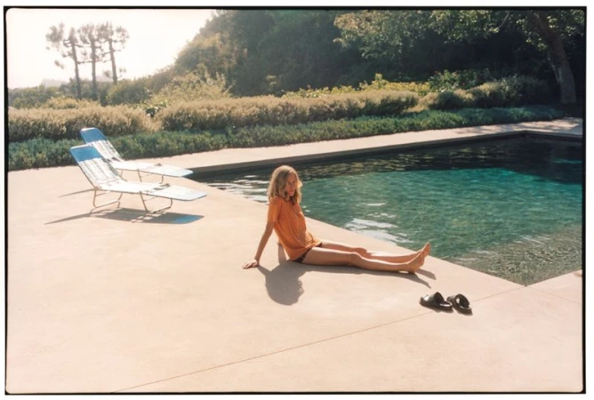 A woman in an orange t-shirt sits poolside on concrete paving next to an outdoor swimming pool and sun loungers.