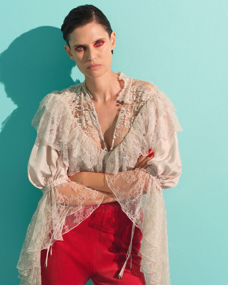 Bianca Balti wearing a ruffled sheer lace blouse and red trousers against a vibrant teal background.