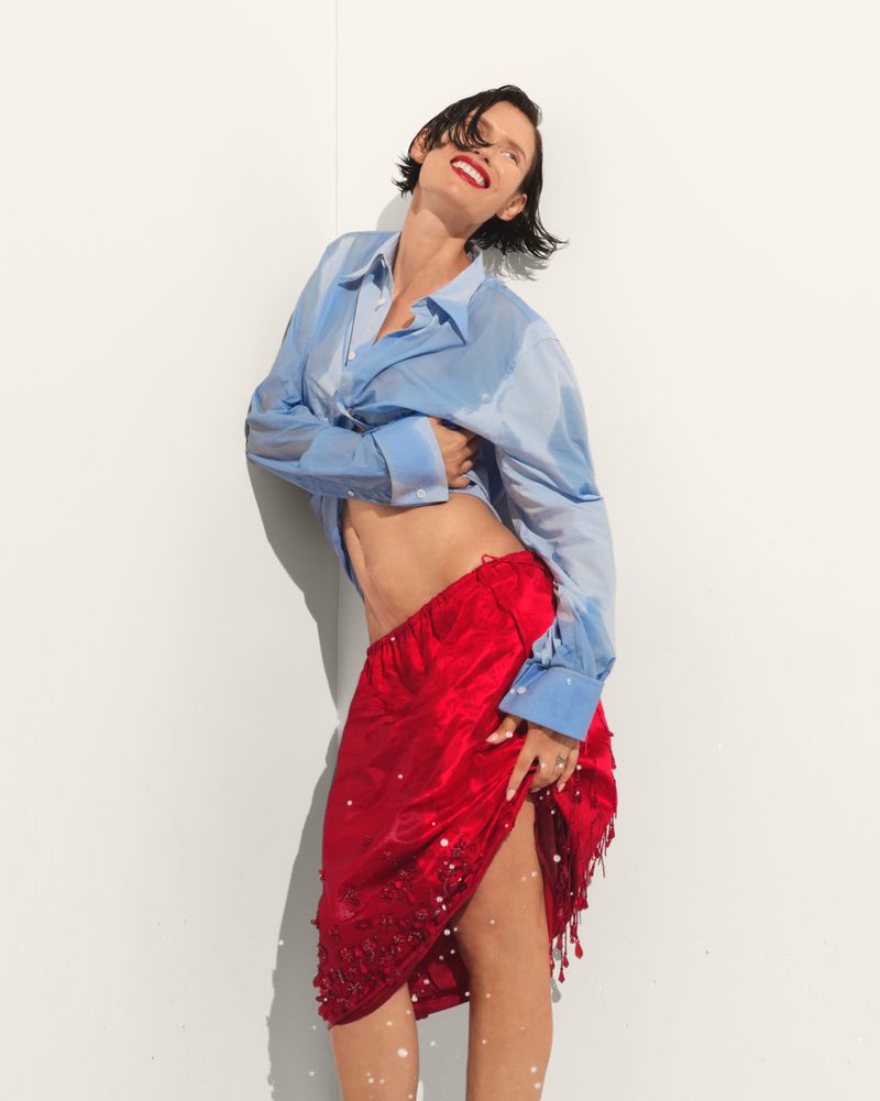Model Bianca Balti wearing a wet blue button-down shirt and red skirt against a white wall for Harper's Bazaar Turkey.