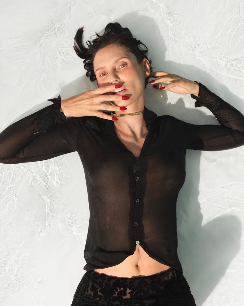 Bianca Balti lying down wearing a sheer black button-up shirt with red nail polish and a gold choker necklace.