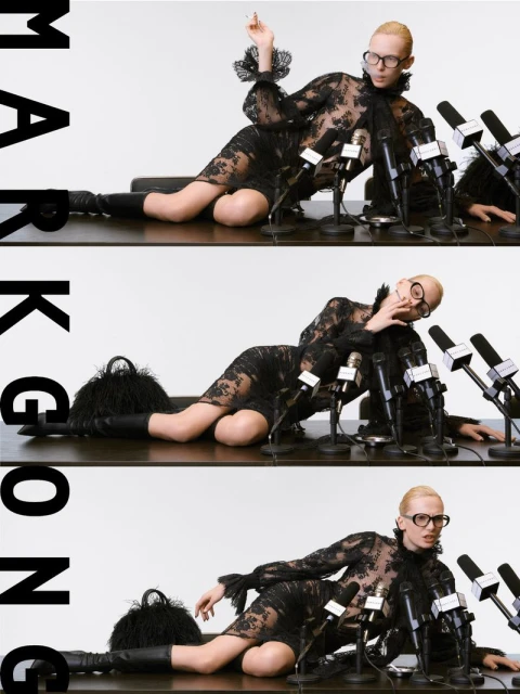 A model in a black lace dress lies on a table covered with microphones in a studio environment, exuding a retro fashion style.