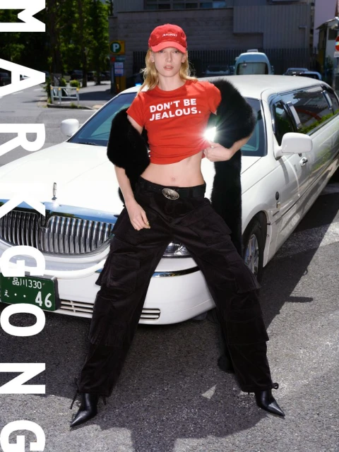 Fashion-forward woman in red top with 'Don't Be Jealous' text, poses with a white limousine on an urban street, blending style and city life.