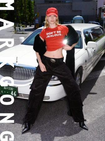 Fashion-forward woman in red top with 'Don't Be Jealous' text, poses with a white limousine on an urban street, blending style and city life.