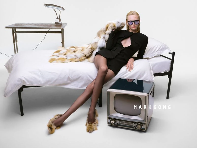 Stylish blonde woman posing on bed with fur coat, sunglasses, black dress, pantyhose, and fur slippers in a white room.