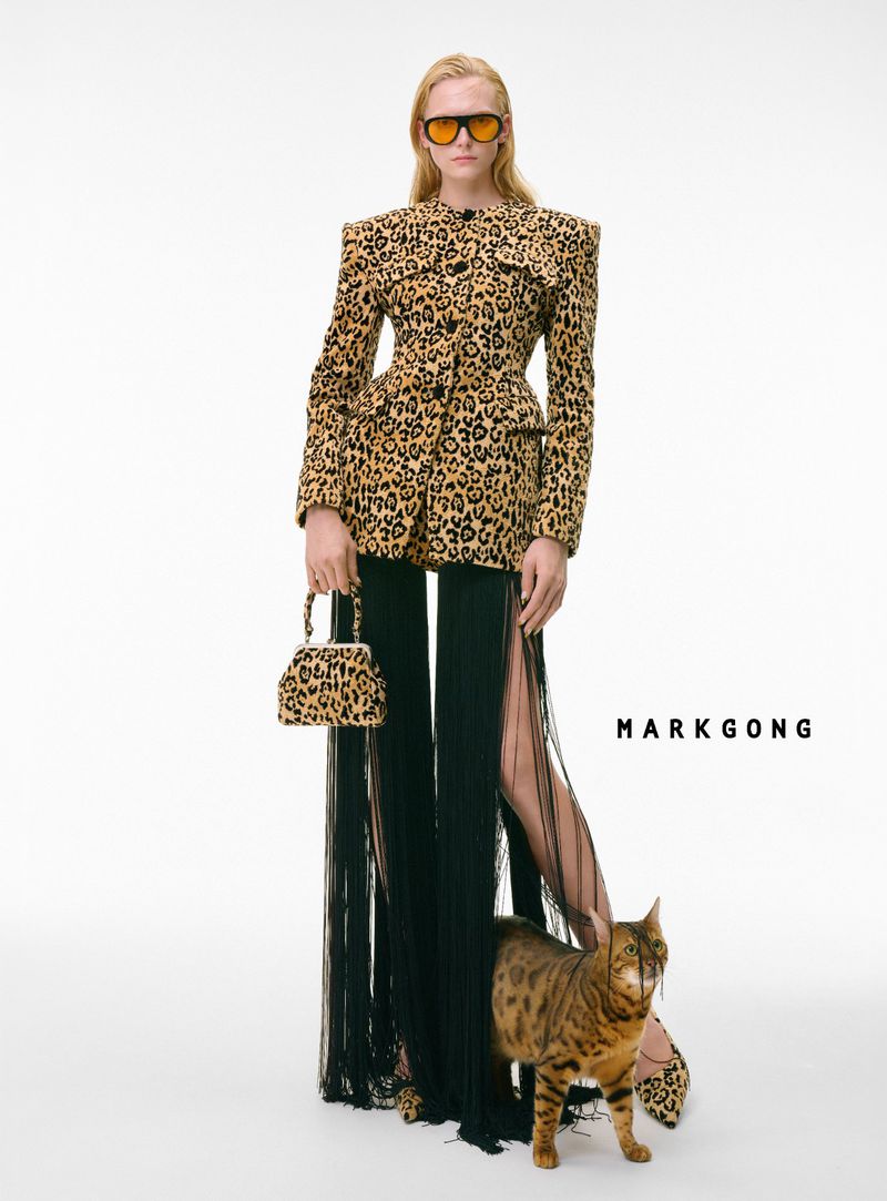 Fashion model poses with a spotted cat, wearing a leopard print jacket, black pants with fringe, and orange sunglasses in a studio.