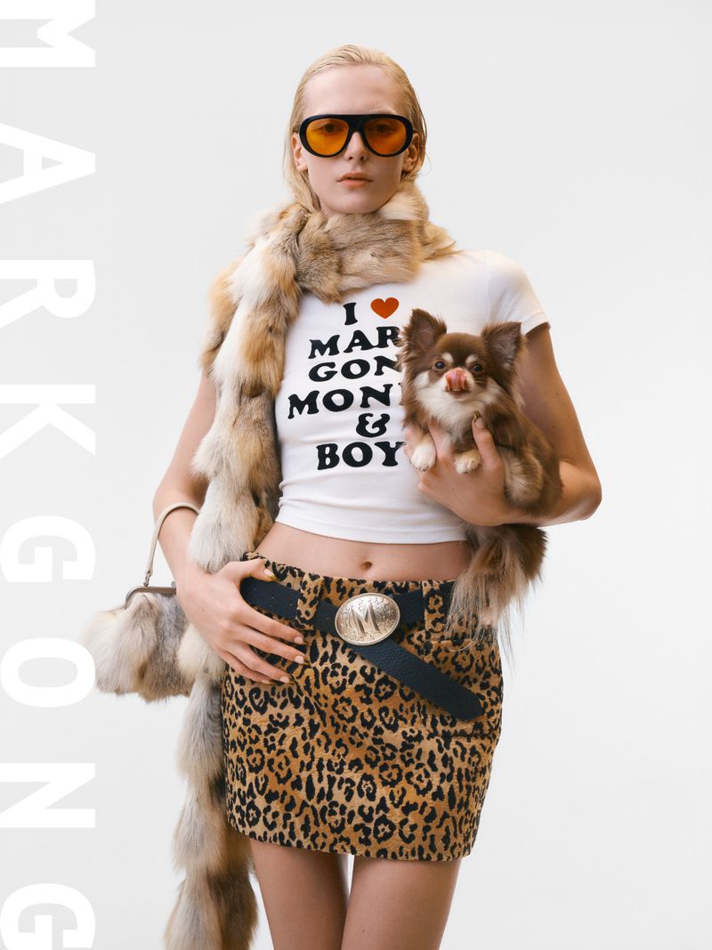 Fashion shot of a female model in a miniskirt and top with a chihuahua, wearing orange glasses and a fur stole against a white backdrop.