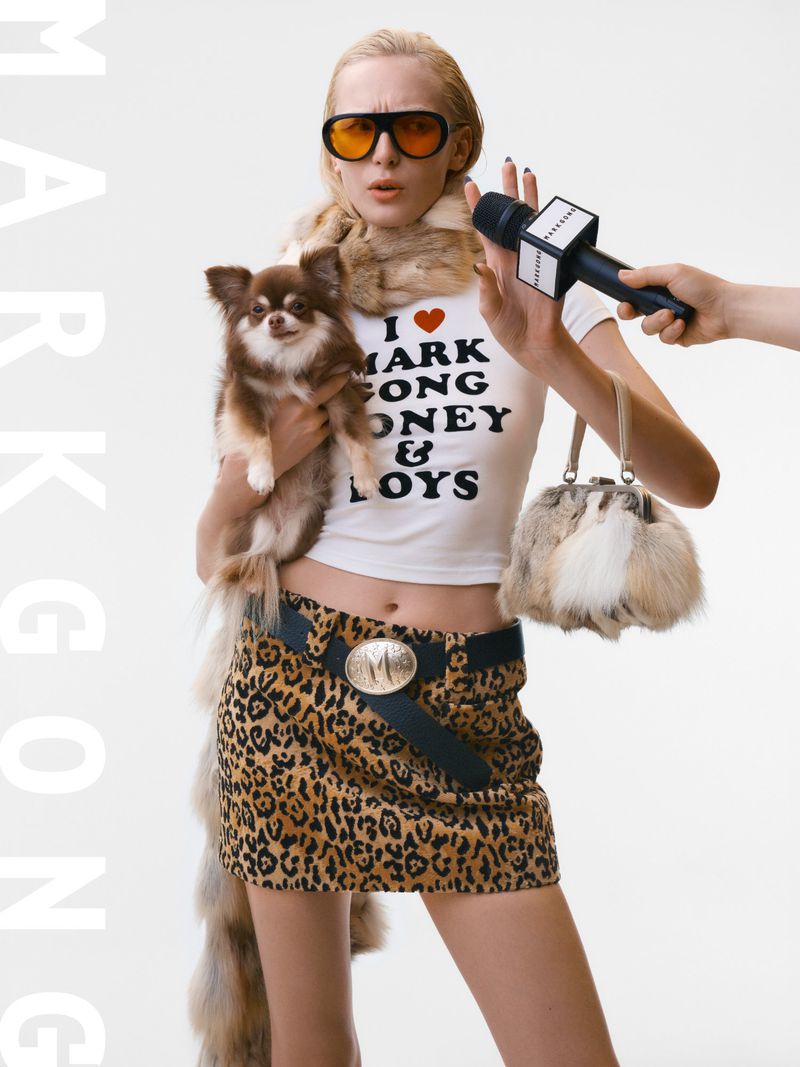 Adult female in leopard-print skirt and fur stole poses with a chihuahua while holding a fur handbag and microphone in a studio.