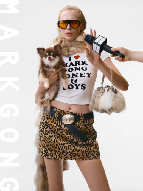 Adult female in leopard-print skirt and fur stole poses with a chihuahua while holding a fur handbag and microphone in a studio.