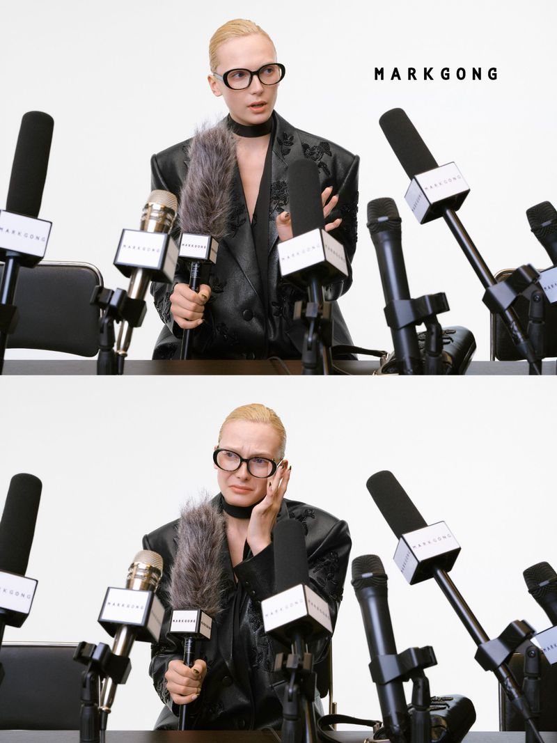 Blonde woman in a black blazer at a press conference style setup, surrounded by microphones, in a brightly lit indoor setting.