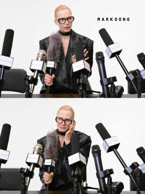 Blonde woman in a black blazer at a press conference style setup, surrounded by microphones, in a brightly lit indoor setting.