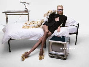 Model Alex Consani wearing a black cutout blazer and tinted glasses, lounging on a white metal bed next to a vintage television.