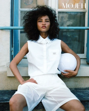 Model with curly afro hair in a sleeveless white collared shirt and athletic shorts holding a white soccer ball outdoors.