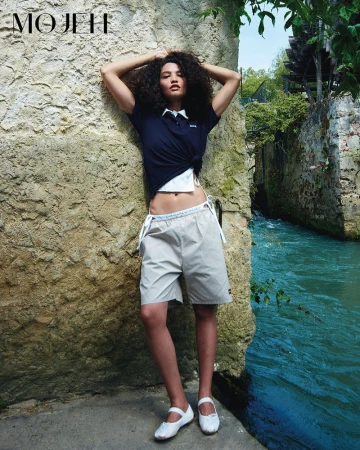 Model posing against a rustic stone wall next to a turquoise river wearing a navy polo shirt, grey bermuda shorts, and white flats.