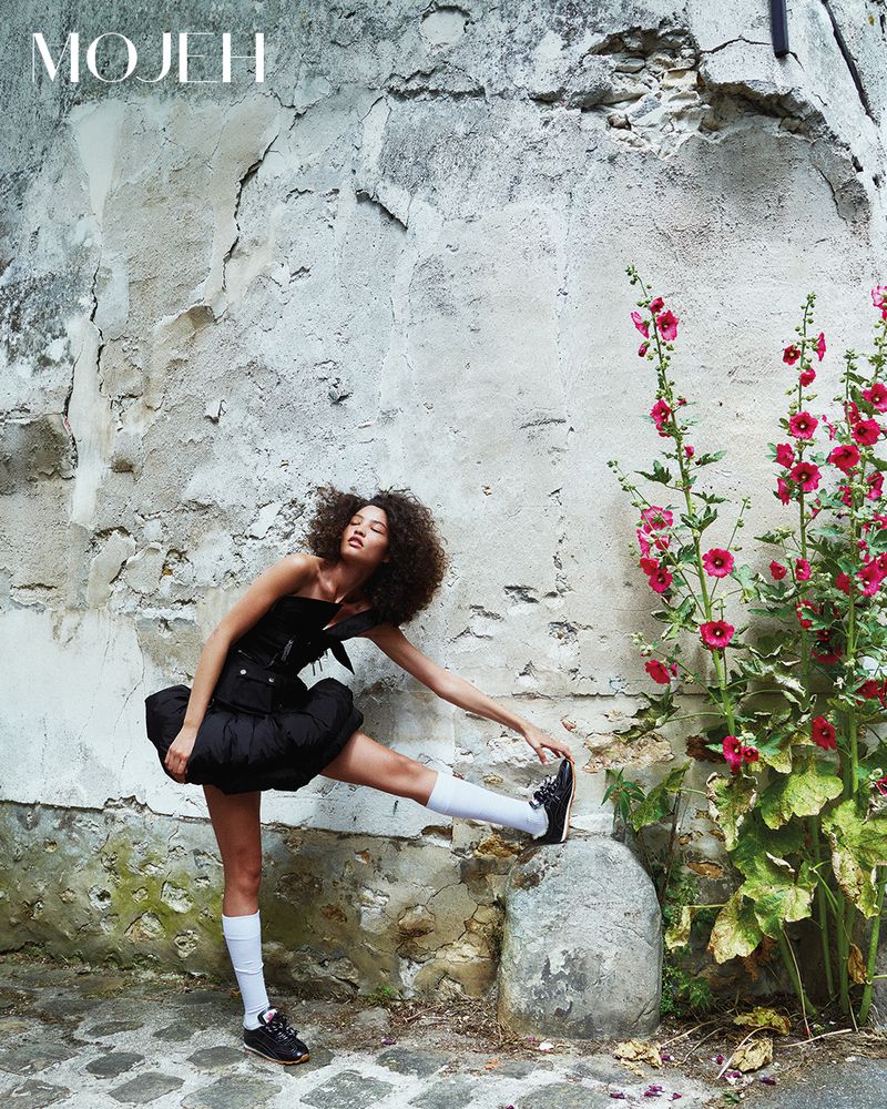 A woman in a black bubble mini dress and white knee-high socks stretches her leg on a stone post by a rustic wall with pink flowers.