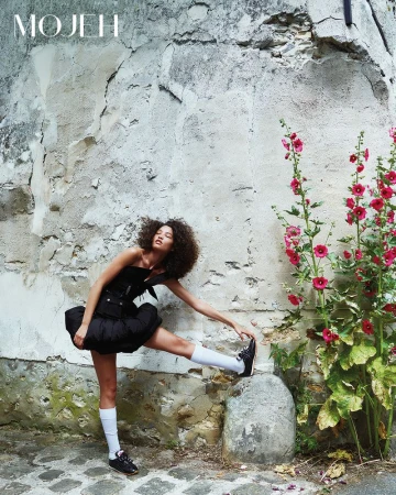 A woman in a black bubble mini dress and white knee-high socks stretches her leg on a stone post by a rustic wall with pink flowers.