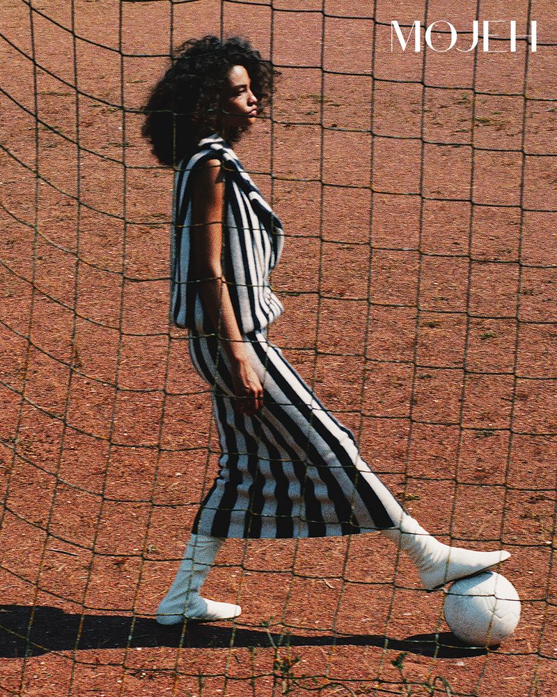 A woman in a black-and-white striped knit outfit and white boots rests her foot on a soccer ball behind a goal net.