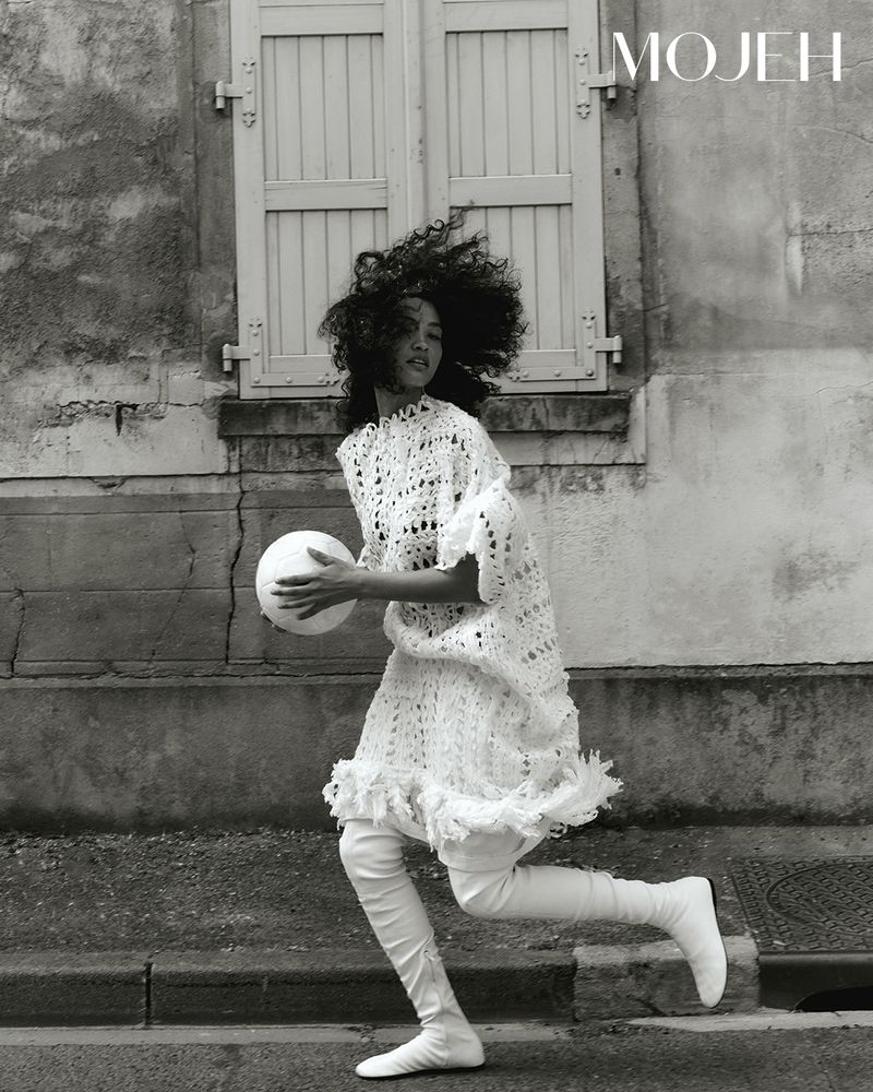 Woman with curly hair in a white crochet dress and tall white boots running outdoors while holding a ball.