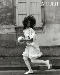 Woman with curly hair in a white crochet dress and tall white boots running outdoors while holding a ball.