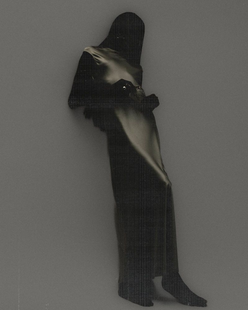 Model silhouetted in a dark hooded garment wearing a shimmering metallic satin maxi dress against a muted grey background.