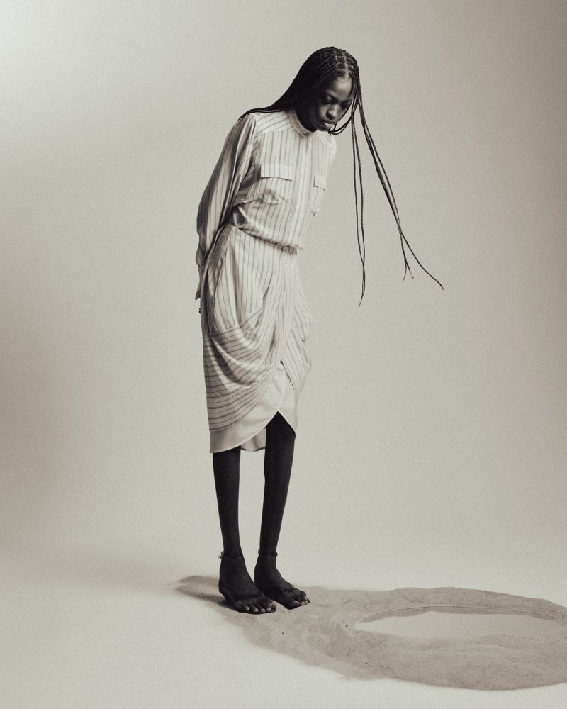 Model J'Adore Benjamin wearing a draped striped dress and black tights, standing near a circle of sand on a seamless studio backdrop.