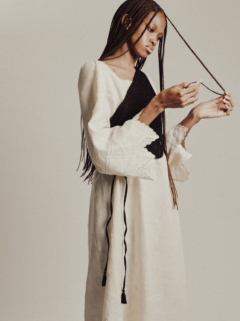 Model J'Adore Benjamin holding her long braids while wearing a cream embroidered dress with black sash against a neutral backdrop.