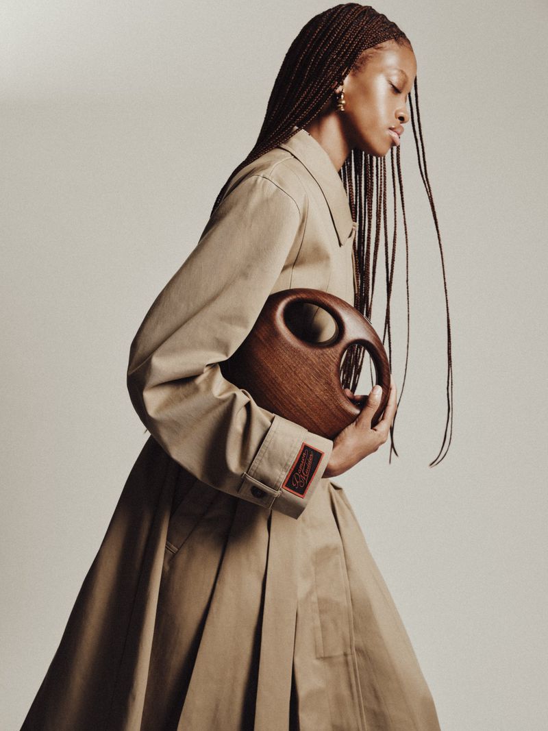 Black woman with long braided hair wearing a beige trench coat and holding a sculpted round wooden handbag against a light backdrop.
