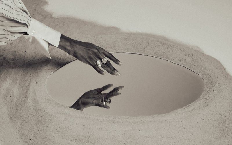 Dark-skinned hand wearing round silver rings reaching toward an oval mirror embedded in fine sand with reflection.