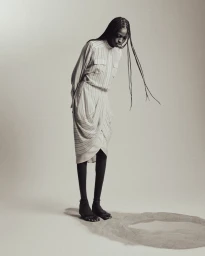 Model J'Adore Benjamin wearing a draped striped dress and black tights, standing near a circle of sand on a seamless studio backdrop.