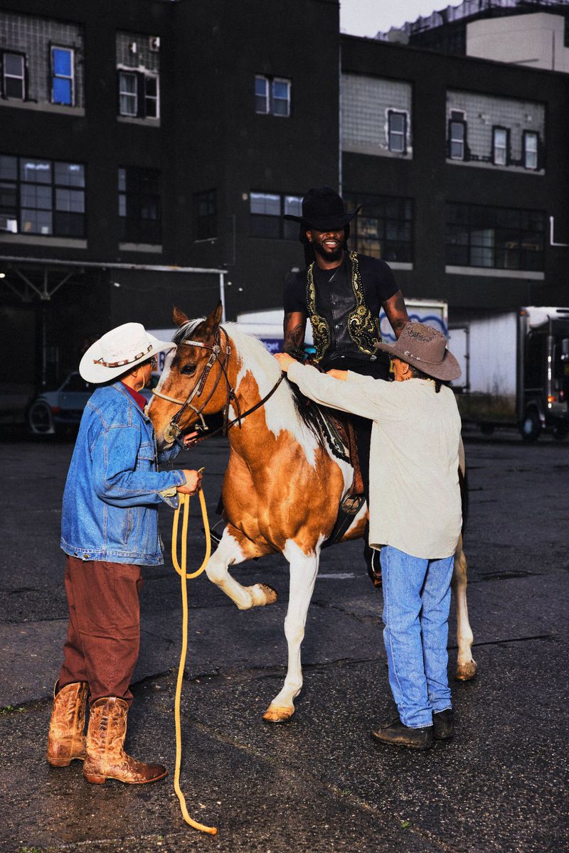 Shaboozey in a black cowboy hat sits atop a brown and white horse assisted by two handlers in an urban asphalt lot.