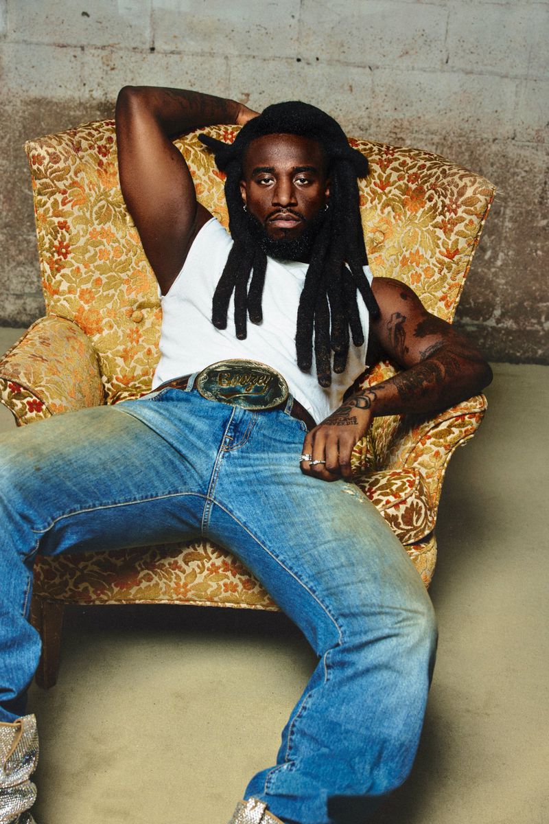 Musician Shaboozey wearing a white tank top and blue jeans, seated in a patterned armchair against a concrete wall.