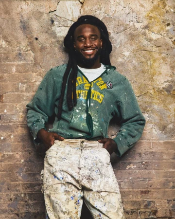 Musician Shaboozey smiling in a paint-splattered green hoodie and cream pants against a rustic brick wall.