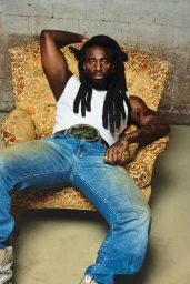 Musician Shaboozey wearing a white tank top and blue jeans, seated in a patterned armchair against a concrete wall.