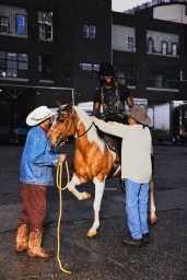 Shaboozey in a black cowboy hat sits atop a brown and white horse assisted by two handlers in an urban asphalt lot.