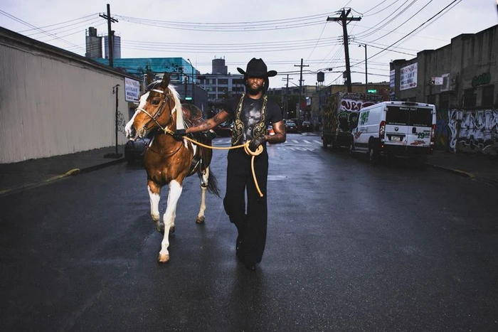 Musician Shaboozey wearing a black cowboy hat walks down a city street leading a brown and white horse by a yellow rope.