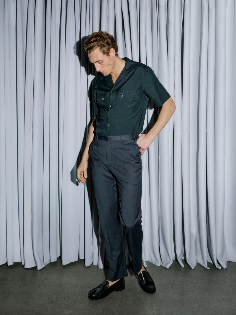 Model Aubrey O'Mahony standing in a dark short-sleeve shirt, pleated trousers, and loafers in front of a grey curtain.