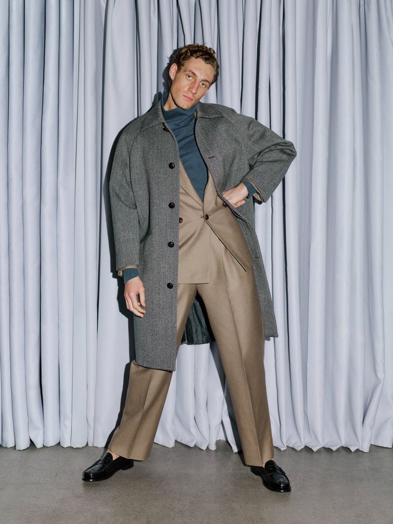 Aubrey O'Mahony posing in a grey overcoat, tan suit, and blue turtleneck against pleated curtains for Les Hommes Publics.