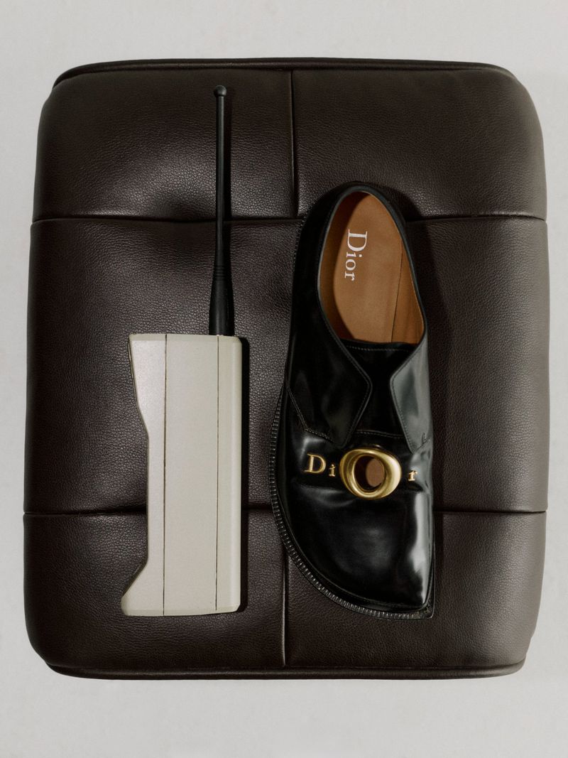 A black leather Dior slip-on shoe with gold hardware and a vintage communication device on a dark brown leather ottoman.